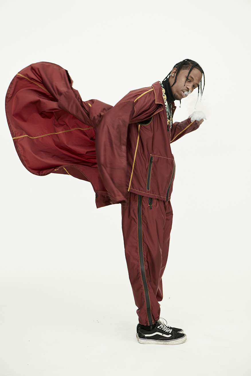 Man poses in a maroon tracksuit, black and white sneakers against a plain white backdrop. The jacket is airborne, creating a dynamic shape.