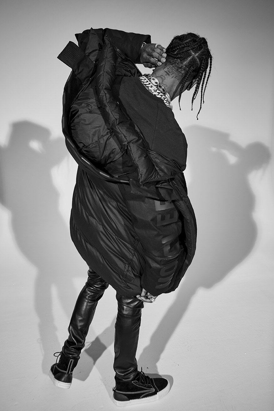 Monochrome portrait of adult male celebrity with braids, wearing leather pants, oversized jacket and platform sneakers in a studio setting.
