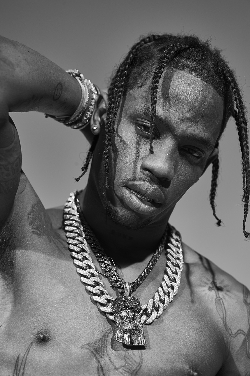 Black and white portrait of a man with braids, wearing heavy jewelry, and posing shirtless against a plain backdrop.