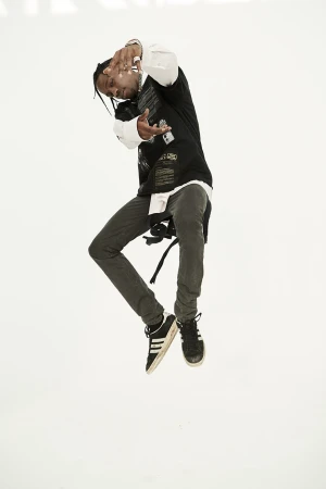 Travis Scott mid-air jumping against a plain white background wearing dark clothing and sneakers