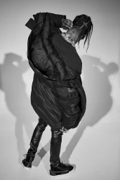 Travis Scott wearing a black puffer jacket and leather trousers viewed from behind against a plain studio background.