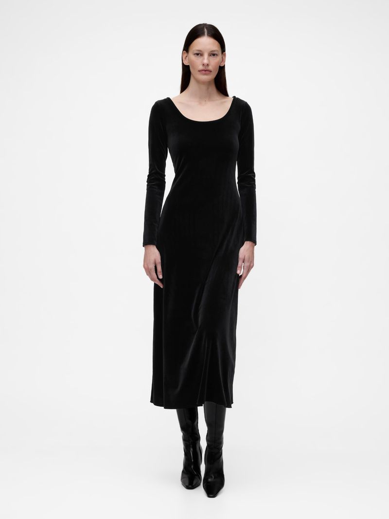 A woman stands in a photo studio wearing a long-sleeved black dress and matching boots against a white backdrop, creating a minimalist fashion image.