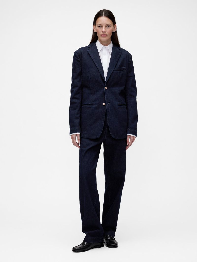 Full-length shot of an adult female with straight brown hair, wearing a navy blue blazer, white shirt, and pants against a white backdrop.