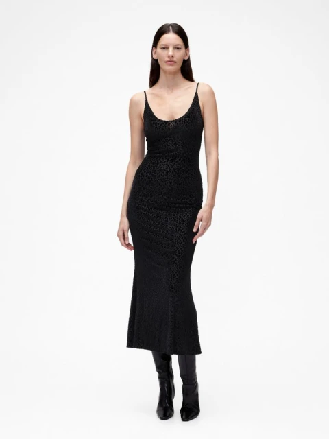 A slender woman poses in a black slip dress and knee-high boots against a white background, showcasing a minimalist studio fashion shot.