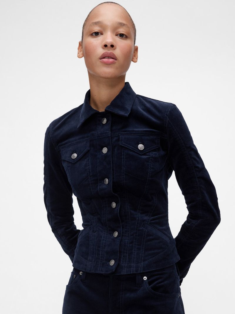 Headshot of a young adult woman modeling a dark blue, button-down, long-sleeved denim jacket with matching denim skirt against a white backdrop.