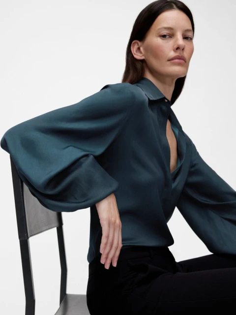 Elegant adult woman in teal satin blouse with bell sleeves sitting on a chair in a studio, looking at the camera.
