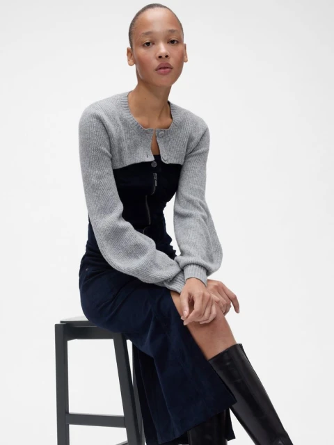 Woman posing in a grey shrug and navy dress, seated on a black stool.  Indoor fashion shot on a white background.  Fashion and style.