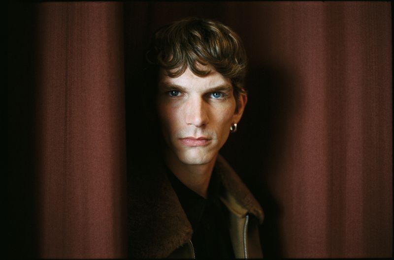 Close-up portrait of a fair-skinned adult man with wavy brown hair and blue eyes, wearing a jacket, posing against a dark red curtain backdrop.