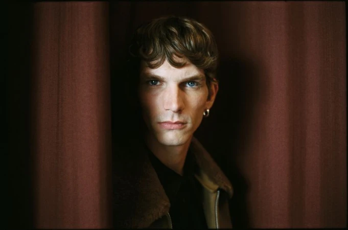 Close-up portrait of a fair-skinned adult man with wavy brown hair and blue eyes, wearing a jacket, posing against a dark red curtain backdrop.