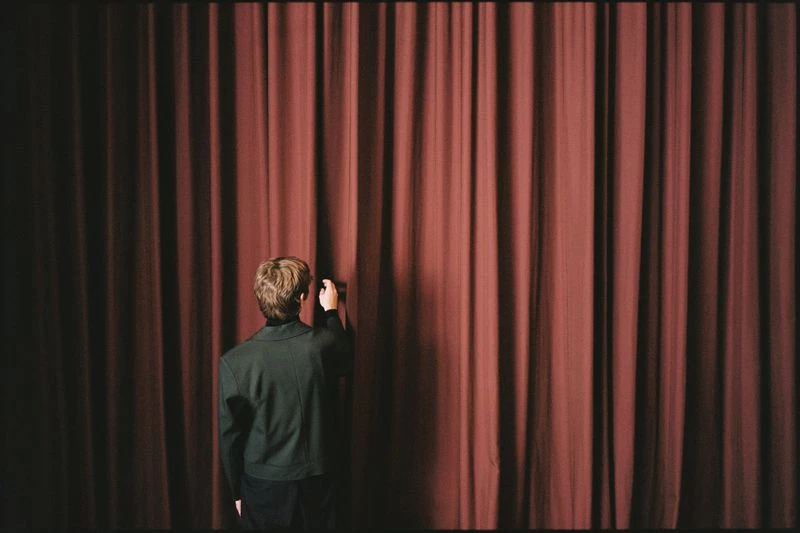 Person in a dark green blazer stands before a reddish-brown curtain in a dimly lit space, pulling the curtain back.