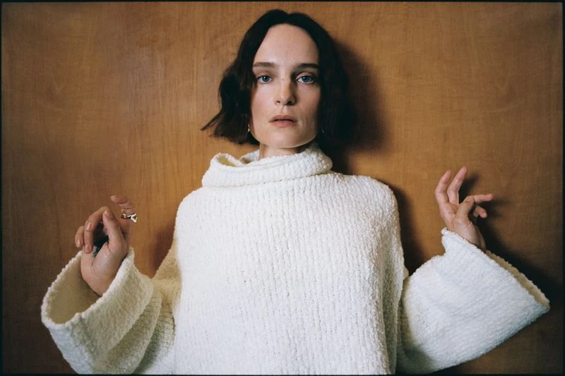 Adult woman posing in white, textured, high-neck sweater against a wood panel background in a portrait shot with natural lighting.