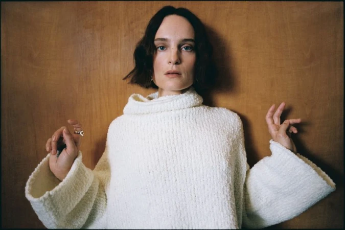 Adult woman posing in white, textured, high-neck sweater against a wood panel background in a portrait shot with natural lighting.