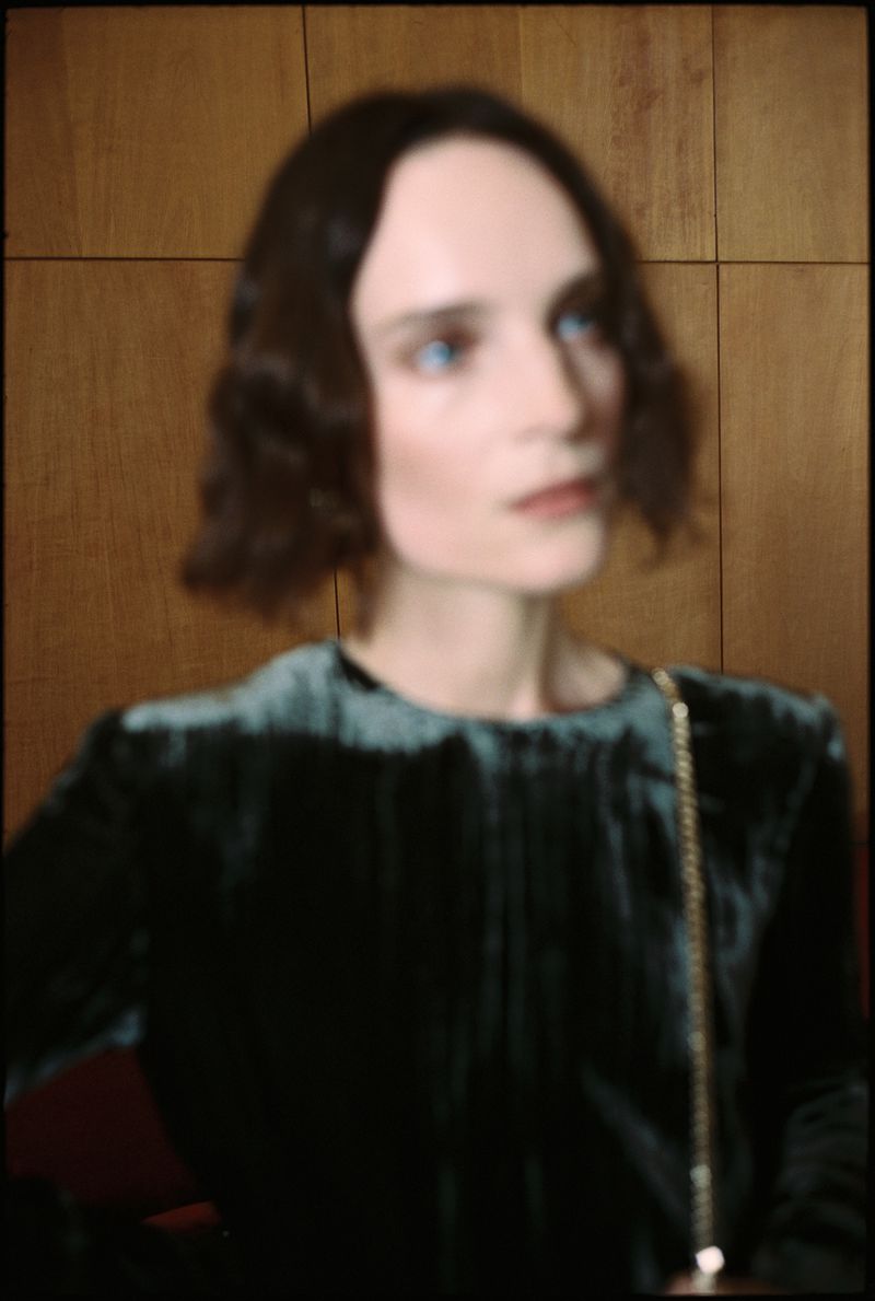 Elegant adult female with short brown wavy hair, wearing a green velvet dress, standing indoors against a brown wooden wall.
