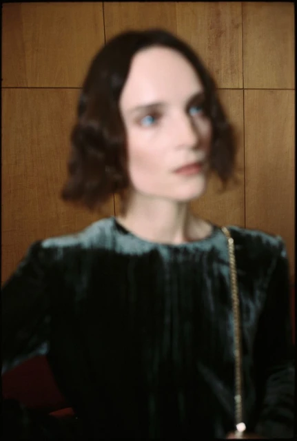 Elegant adult female with short brown wavy hair, wearing a green velvet dress, standing indoors against a brown wooden wall.