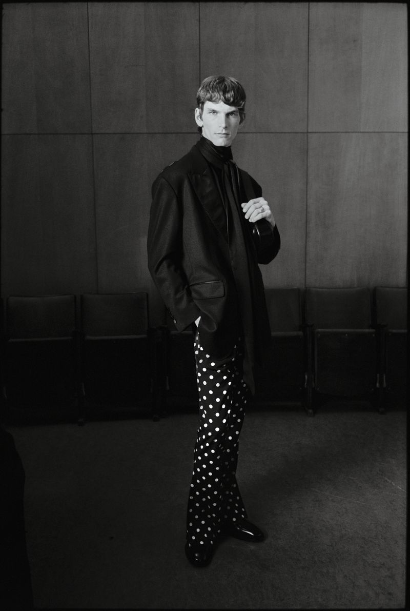 Monochrome portrait of a male model in a suit jacket and polka dot pants standing in front of a grey wall with chairs.