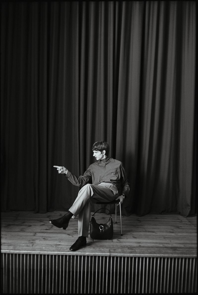 Adult man poses on stage in a relaxed sitting position with one arm extended, wearing a long-sleeve shirt, pants, and ankle boots.