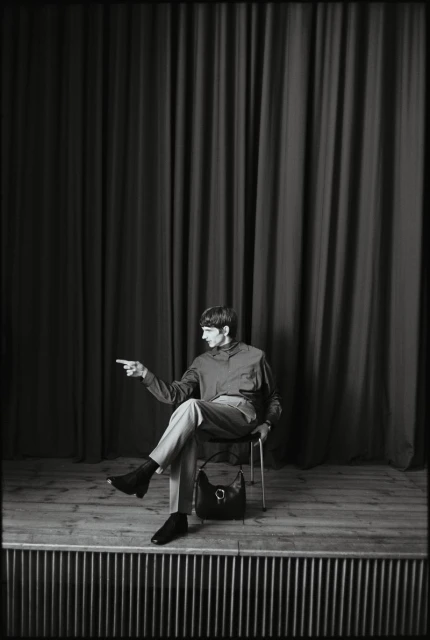 Adult man poses on stage in a relaxed sitting position with one arm extended, wearing a long-sleeve shirt, pants, and ankle boots.
