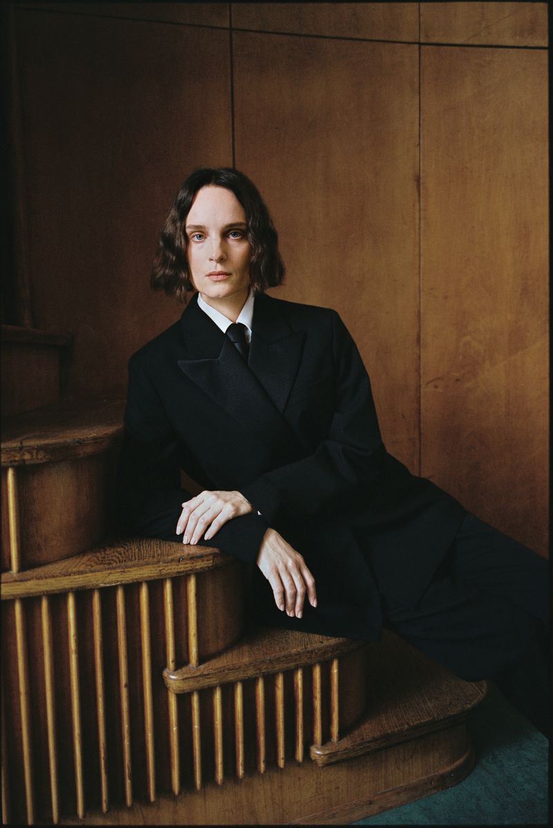 Portrait of a long-haired adult woman wearing a black suit and tie, sitting on wooden stairs against a wooden background indoors.