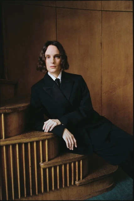 Portrait of a long-haired adult woman wearing a black suit and tie, sitting on wooden stairs against a wooden background indoors.