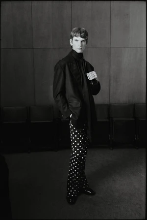 Monochrome fashion portrait of a man in a dark blazer and polka dot trousers standing before panelled walls.