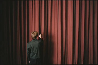 Man in dark jacket seen from behind pulling back heavy red stage curtains in a dramatic theatre setting.