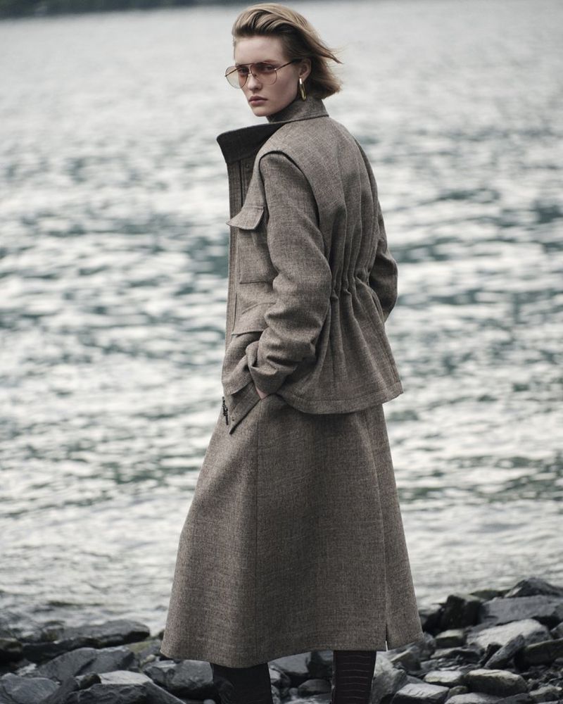 Adult woman in a wool jacket and skirt poses near a body of water on a cloudy day, accessorized with gold earrings and sunglasses.
