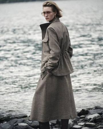 Adult woman in a wool jacket and skirt poses near a body of water on a cloudy day, accessorized with gold earrings and sunglasses.