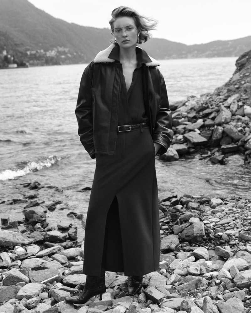 Black and white outdoor shot of a woman in a coat and dress standing on rocks near the water's edge with mountains in the background.