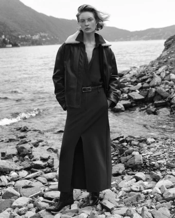 Black and white outdoor shot of a woman in a coat and dress standing on rocks near the water's edge with mountains in the background.