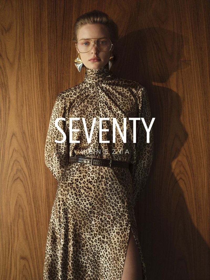 Woman in animal-print dress and aviator glasses poses in front of wood panel. Fashion shot with seventies style.