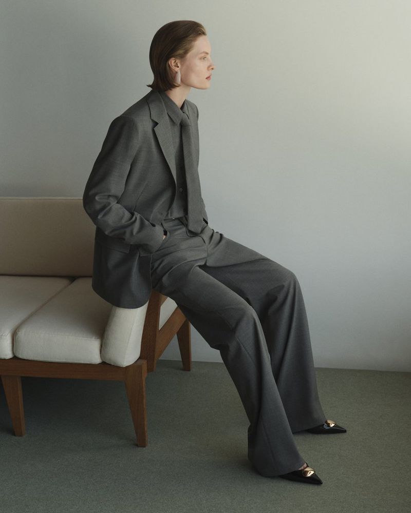 A woman in a grey pantsuit sits on a couch in a photo studio. The image focuses on fashion, style, and modern clothing.