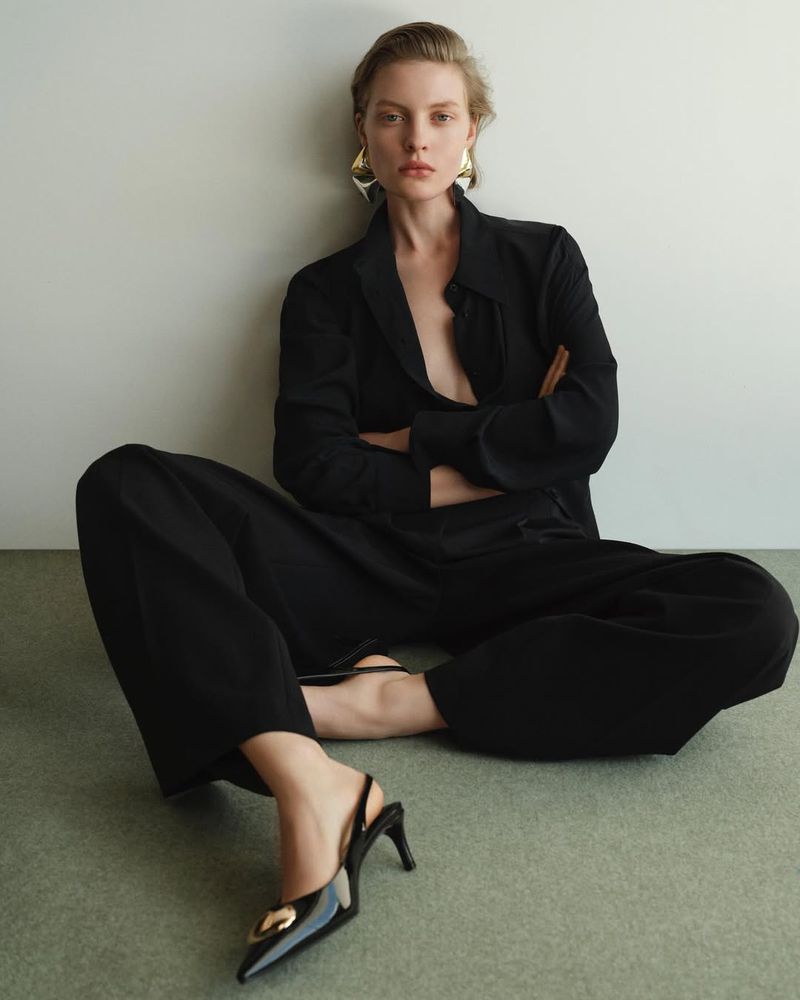 An adult woman sitting on the floor with legs crossed, wearing a black suit jacket, black wide-legged pants and shiny black heels.