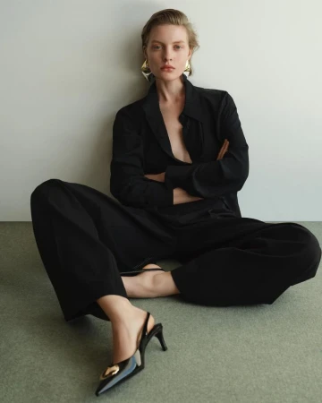 An adult woman sitting on the floor with legs crossed, wearing a black suit jacket, black wide-legged pants and shiny black heels.