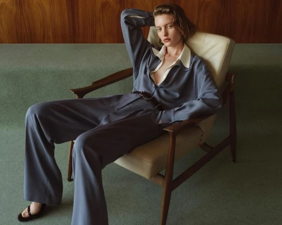 Elegant woman in stylish blue suit and open white shirt reclining in vintage armchair in neutral interior with wood paneling, sandals.