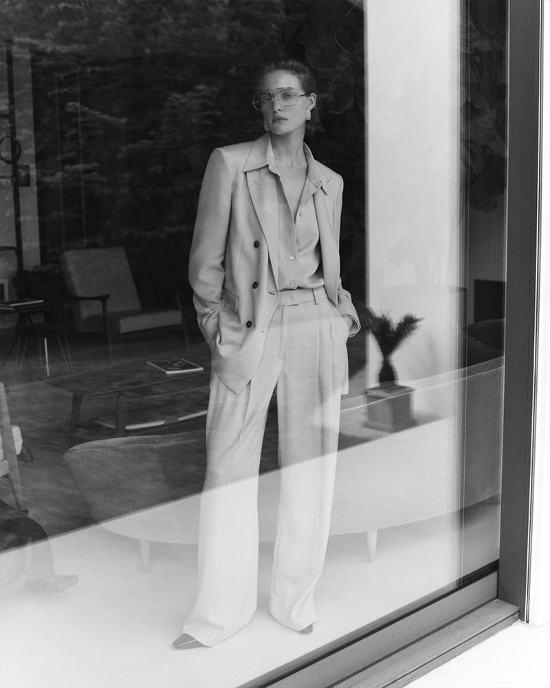 Black and white portrait of an adult woman wearing a suit and eyeglasses, standing indoors, reflected in a window with an outdoor view.