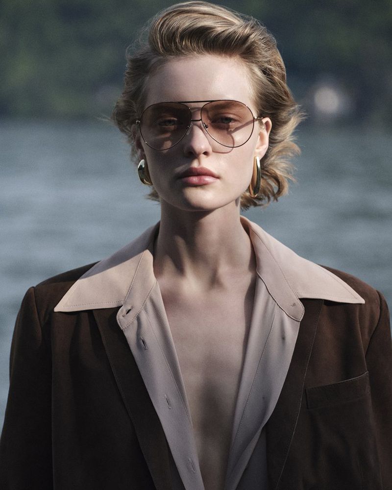 Elegant woman with blond hair and aviator sunglasses poses outdoors in a brown blazer and button-down shirt against a blurred lake backdrop.
