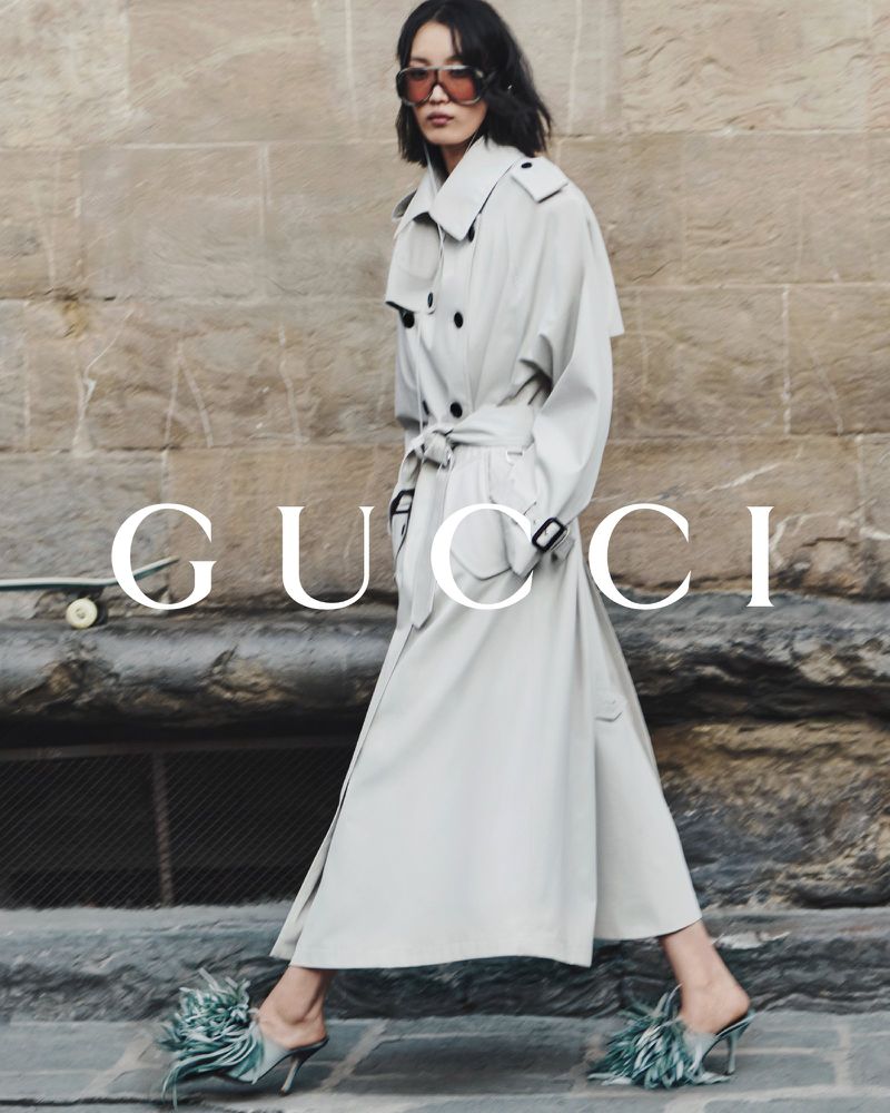 Elegant Asian woman in a trench coat and feathered heels walks against a stone wall with 'Gucci' logo in a fashion editorial.
