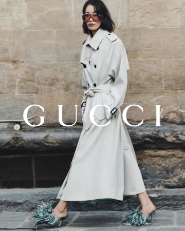 Elegant Asian woman in a trench coat and feathered heels walks against a stone wall with 'Gucci' logo in a fashion editorial.