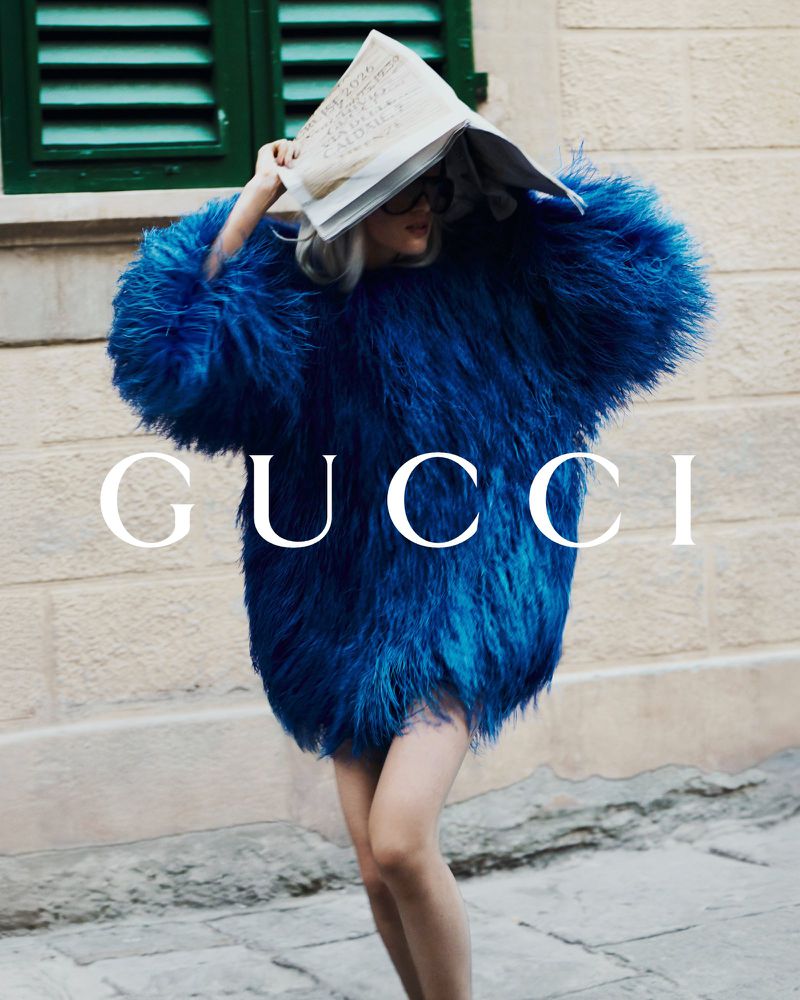 Stylish woman in an oversized blue feather jacket with a newspaper hat, photographed outdoors against a light wall. Fashion editorial.