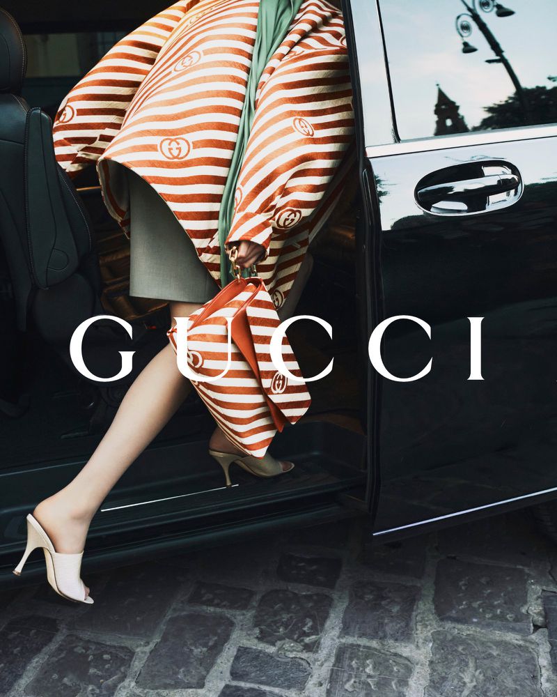 Fashion shot of a woman exiting a black car, wearing a striped jacket and carrying a matching handbag. Gucci logo is visible.