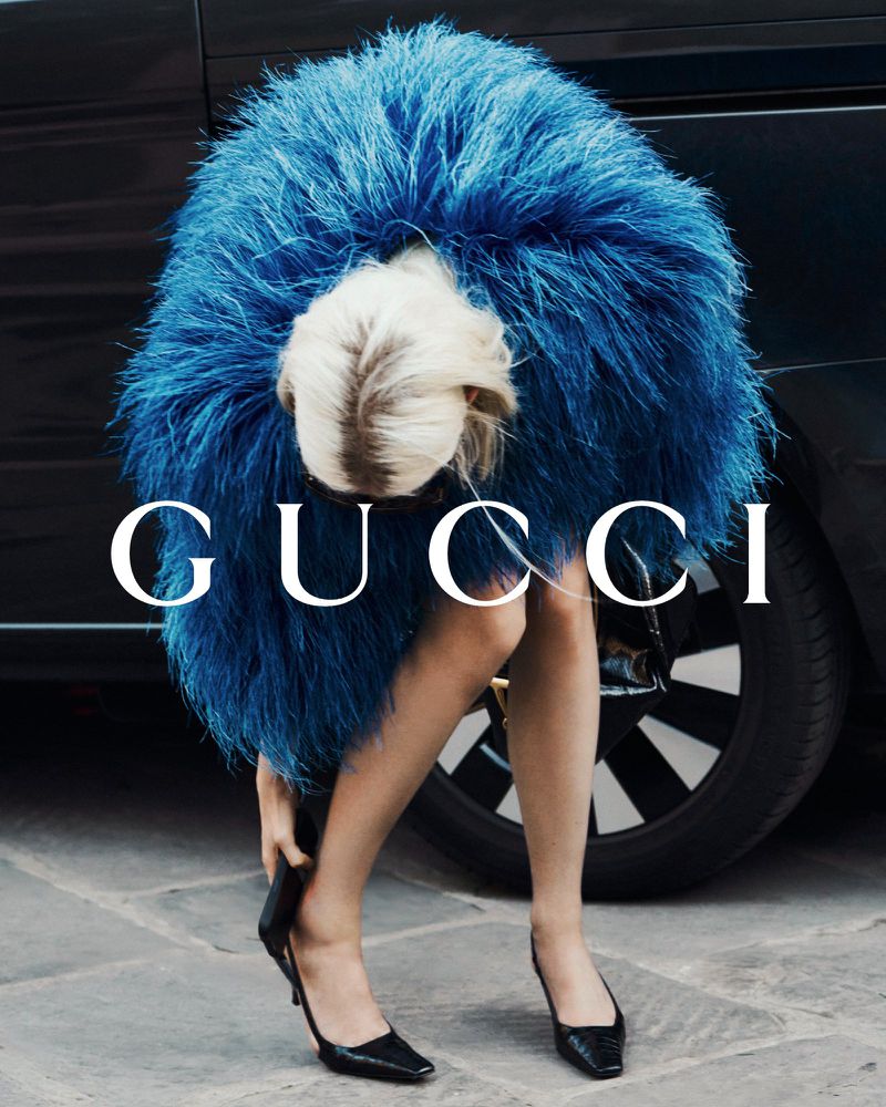A woman in a blue feather coat adjusts her black high heel shoe next to a black car. The 'GUCCI' logo is visible in the foreground.