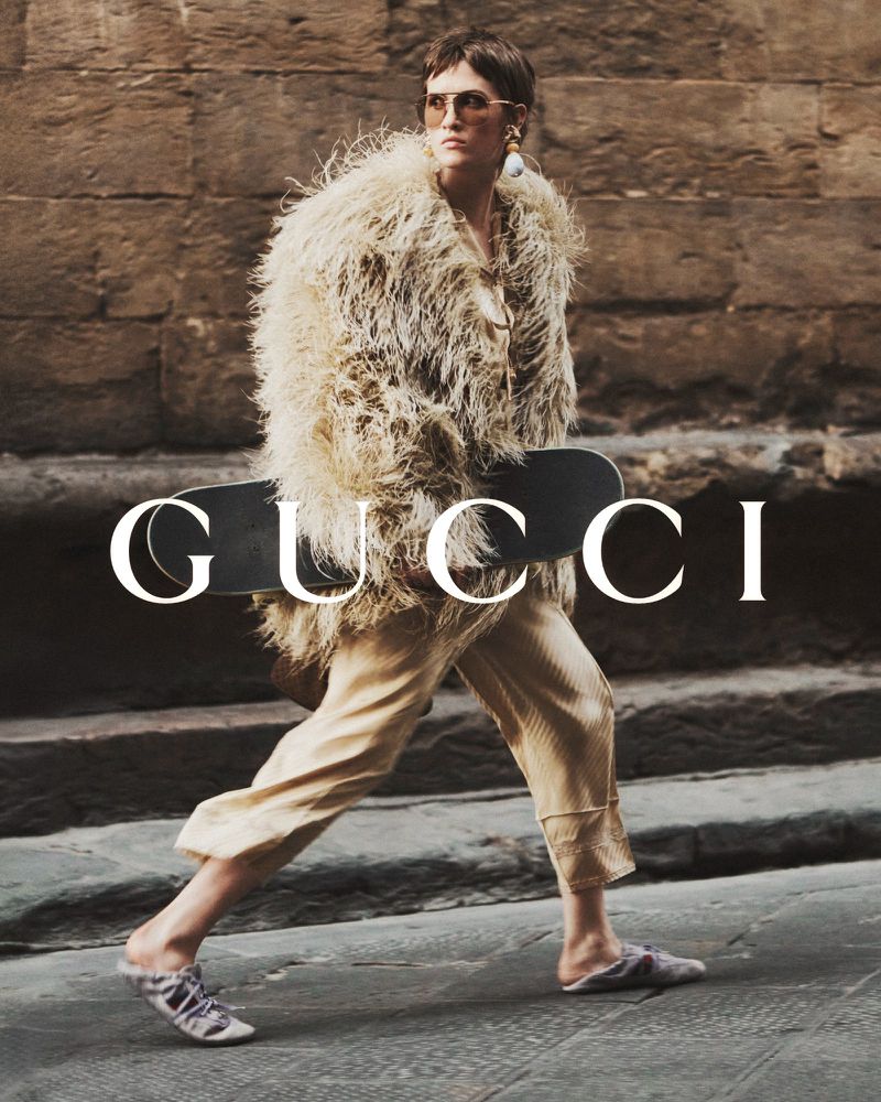 Fashion photo of a woman in a fur coat and sunglasses, holding a skateboard in front of a building with the Gucci logo overlaid.