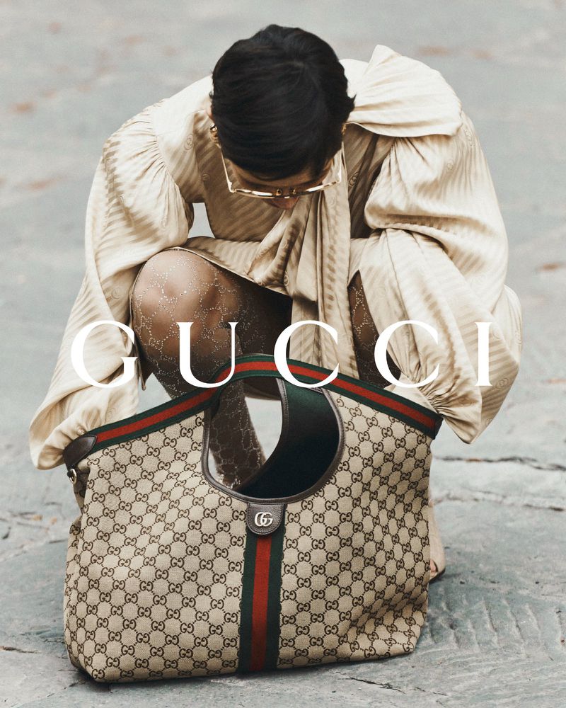 Woman crouches near a Gucci handbag with the Gucci logo visible. She wears a tan colored dress and glasses, fashion advertisement.
