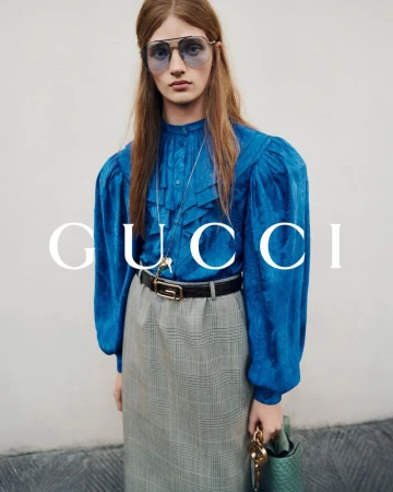 A person with long brown hair, wearing a bright blue blouse, aviator glasses, a long skirt and carrying a handbag, stands in a fashion shot.