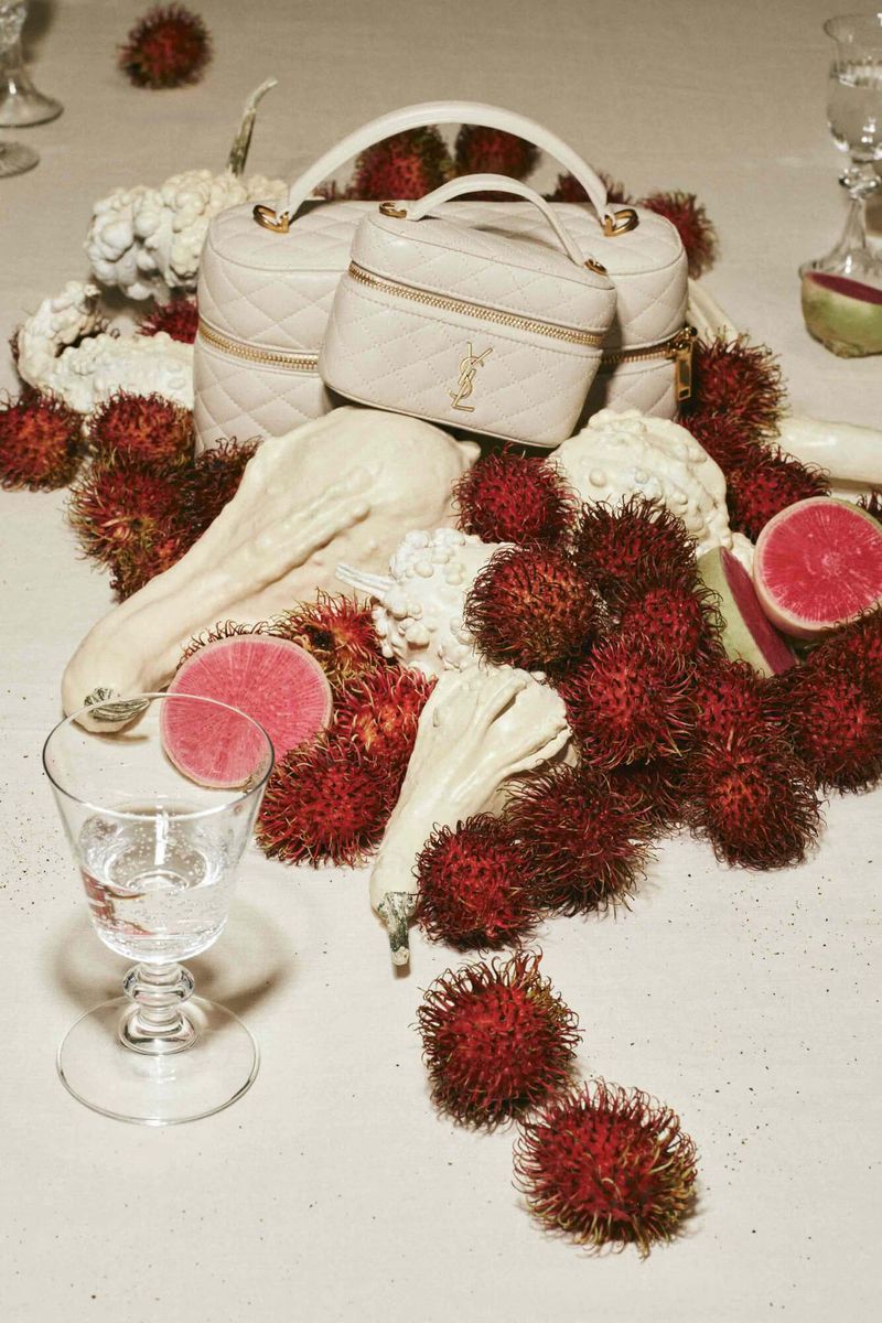 Two YSL beige vanity bags with quilted leather handles sit among lychee fruit, white squash, and water glasses on a beige surface.
