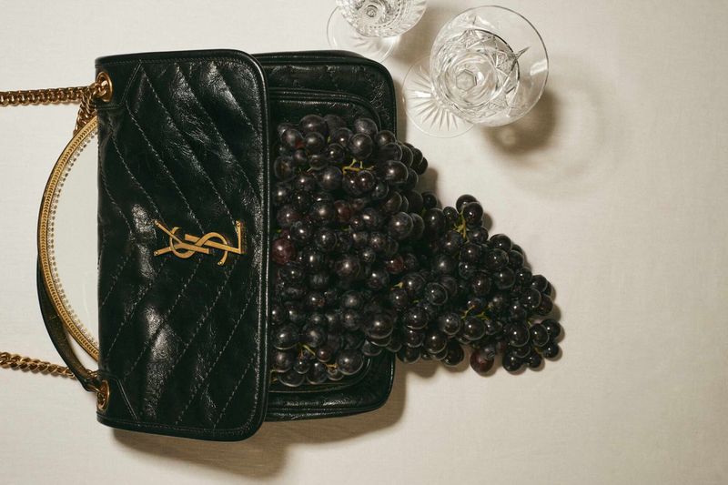 Black leather handbag with golden YSL logo filled with black grapes, crystal glassware on neutral surface, high-angle product shot.