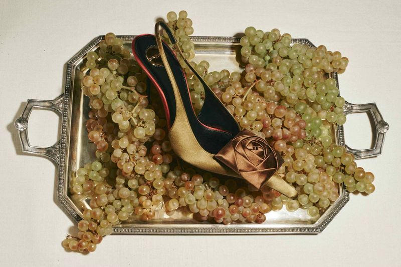 Tan high-heeled shoe with a rose on a silver tray surrounded by green grapes, creating a luxurious and fashionable still life.