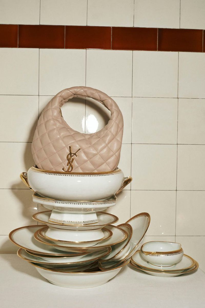 Light pink leather handbag with a gold logo resting atop a tower of white and gold decorative plates against a white tile background.