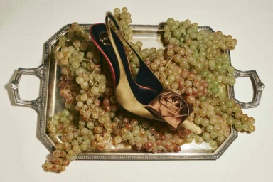 Gold high-heeled slingback shoe resting on a silver tray surrounded by fresh green grapes.