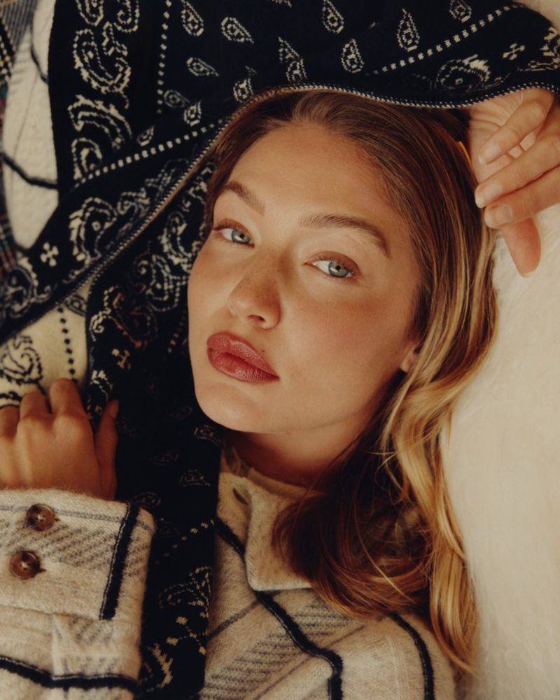 Close-up of a woman with blonde hair and blue eyes wearing a beige plaid coat, looking at the camera with a neutral expression.